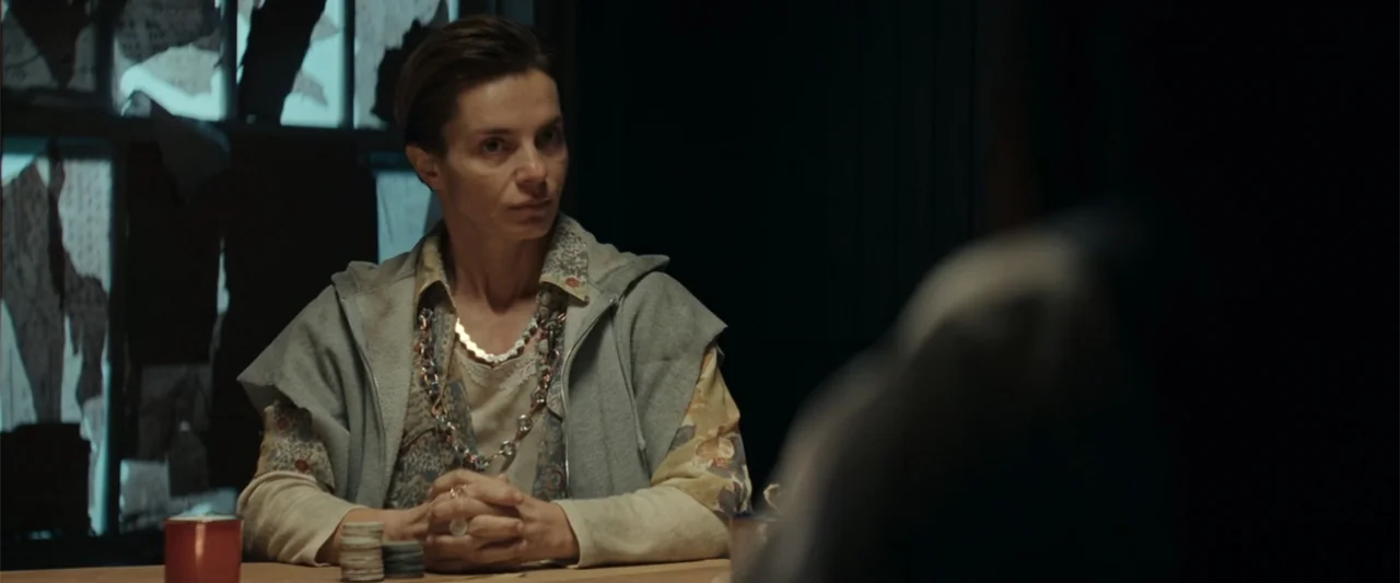 McWalter (2025) movie still: medium — A dimly lit room features a window with torn paper sheets obscuring the glass panes. A woman sits at a wooden…