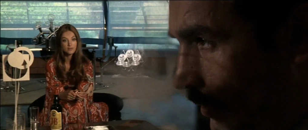 Don’t Torture A Duckling (1972) movie still: medium — A living room interior with modern furniture and large windows overlooking exterior greenery.…