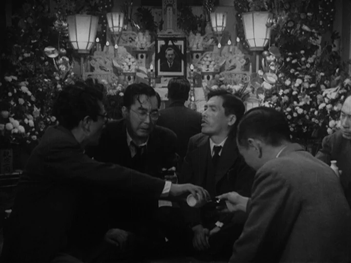 Ikiru (1952) movie still: medium — An interior funeral service features a central framed photograph surrounded by floral arrangements and lanterns.…