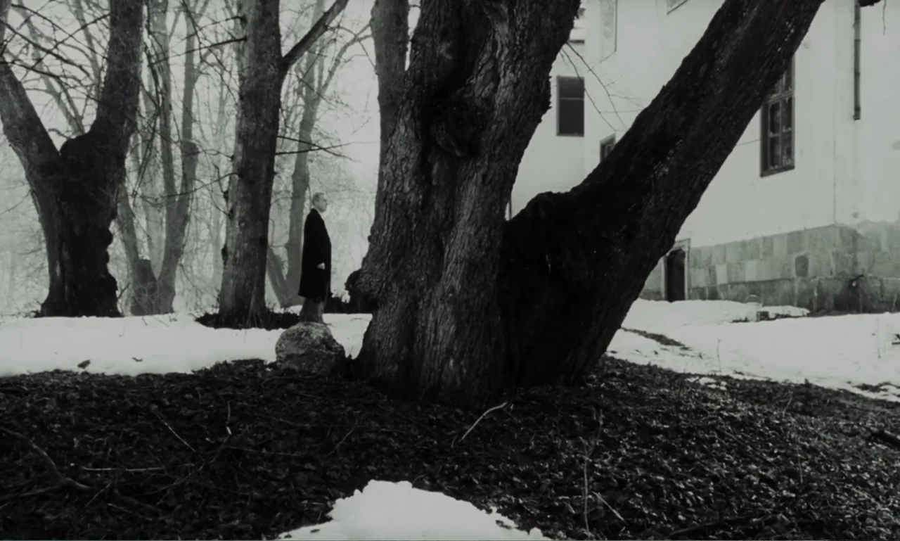 The Sacrifice (2015) movie still: wide — A snowy outdoor area features several large bare trees and a white stone building. An elderly man in a dark…