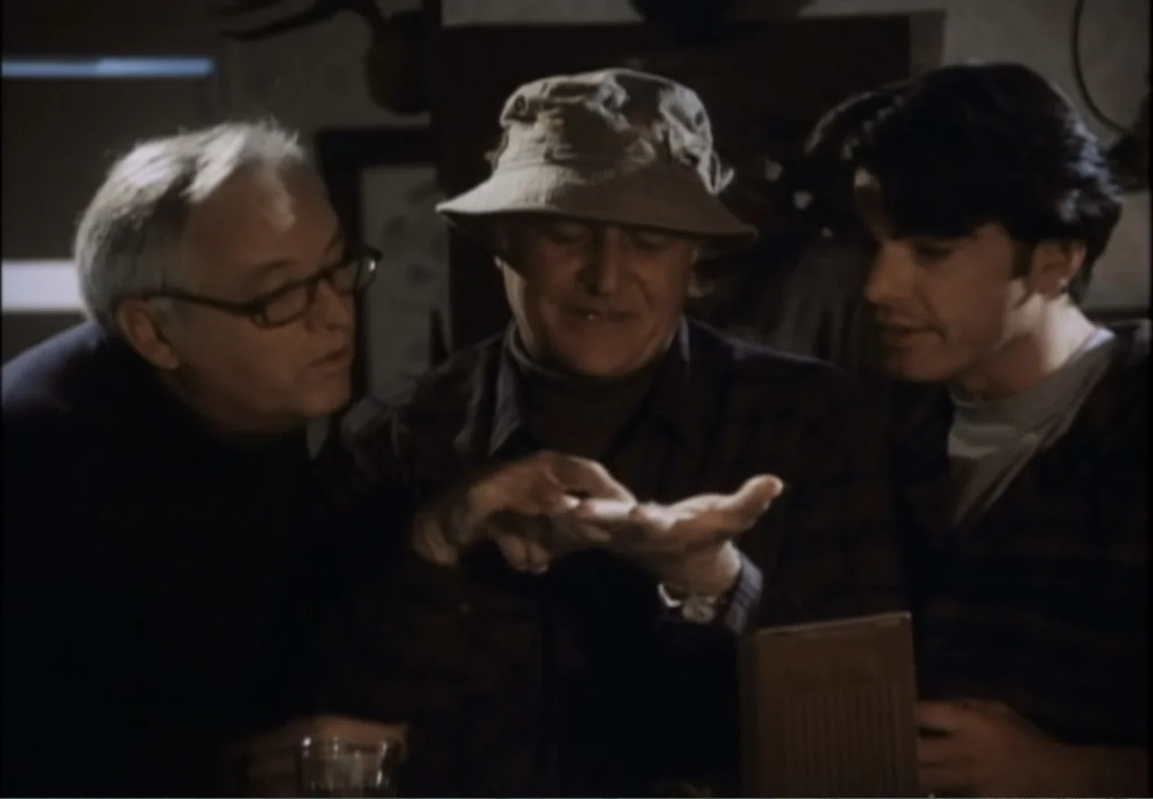 White Mile (1994) movie still: medium — Three men stand clustered in a dark, dimly lit indoor room with rustic decor. The man in the center holds an…