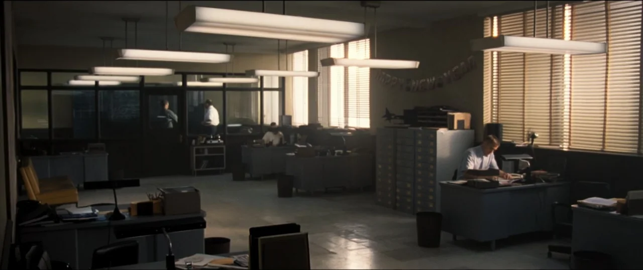 First Man (2018) movie still: wide — An open-plan office features rows of grey desks under long rectangular overhead fluorescent fixtures. One man…