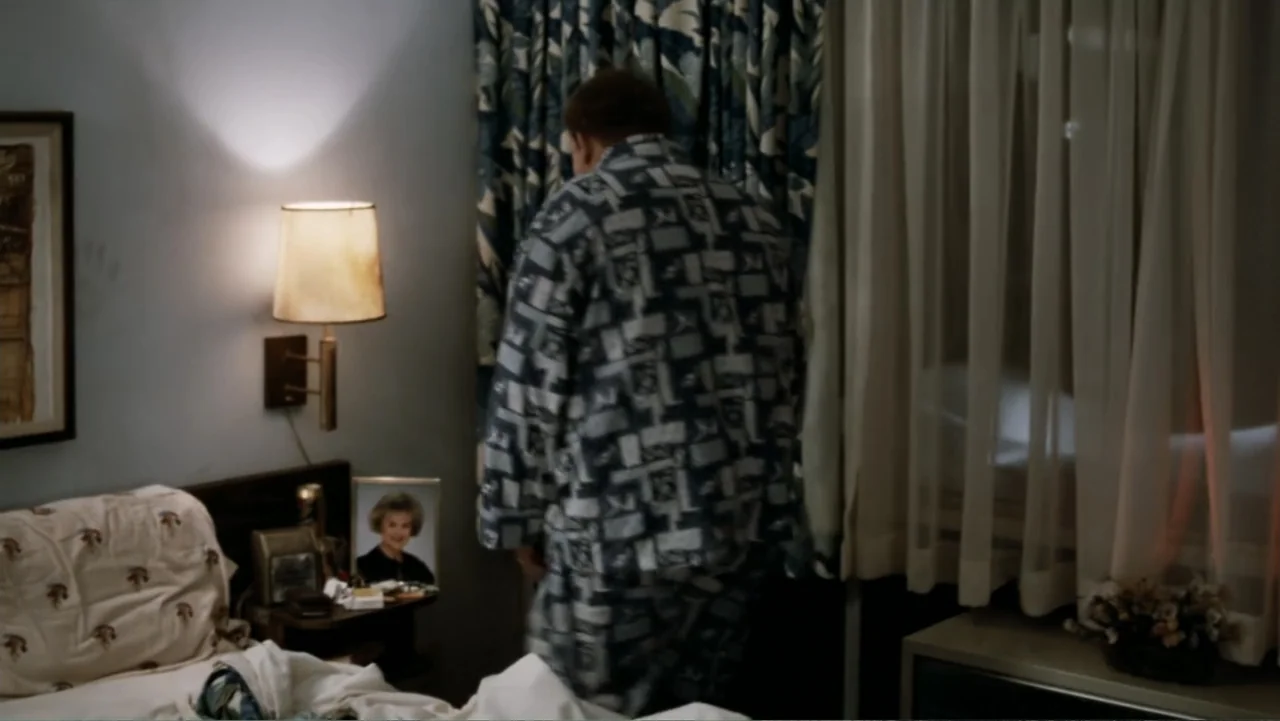 John Candy: I Like Me (2025) movie still: medium — A dimly lit motel room features a bed and a small bedside table. A man in pajamas stands facing…