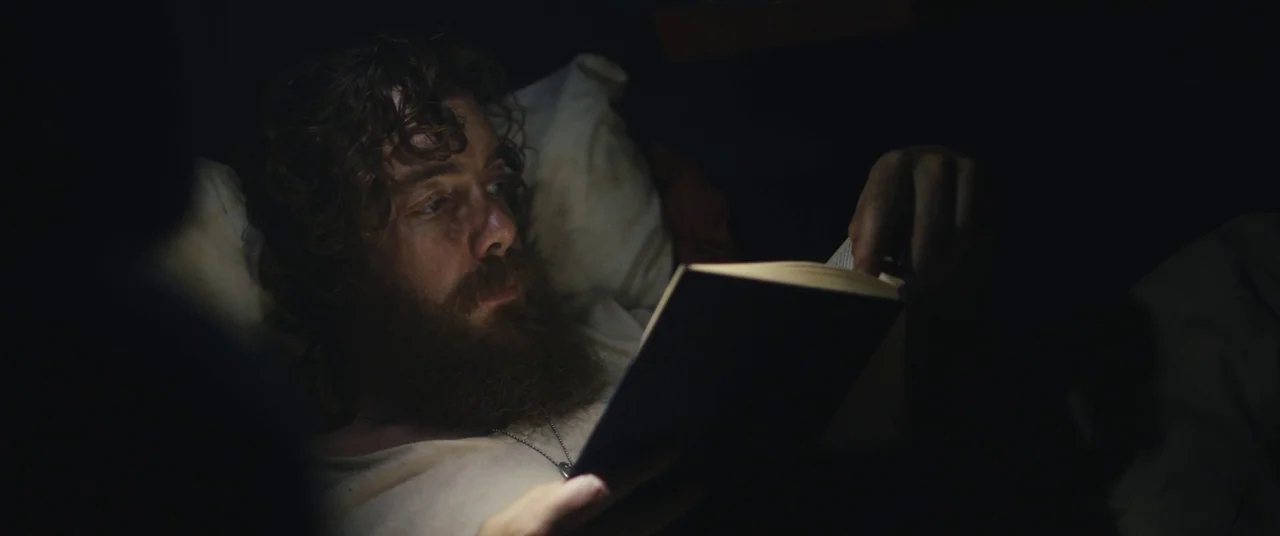 Blue Ruin (2013) movie still: closeup — A man lies in a dark room under soft bed covers. He holds a book while looking at the pages by a light source.