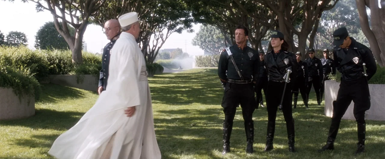 Demolition Man (1993) movie still: fullbody — An outdoor lawn area features a path lined with trees and several uniformed individuals. A man in a…