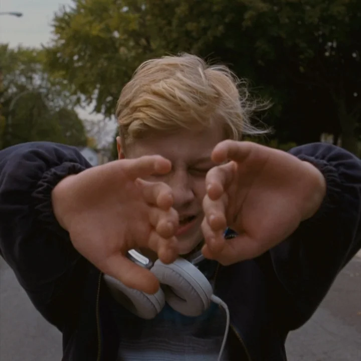Mommy (2014) movie still: closeup — A teenager stands outdoors on an asphalt street with trees visible in the background. The young male subject…
