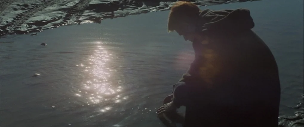 The Man Who Fell To Earth (1976) movie still: medium — A person kneels at the edge of a shallow, muddy pool in a desolate area. The individual…