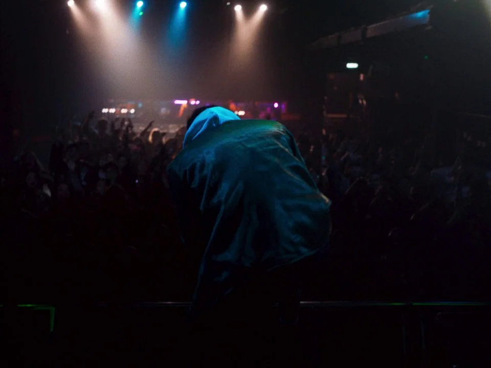 Mogul Mowgli (2020) movie still: medium — A dark concert venue with a large silhouette of a person on a stage. The subject crouches low while facing…
