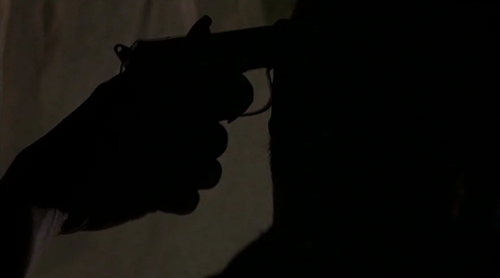 Jackie Brown movie still: closeup — A dark space with heavy shadows concealing the surroundings. A silhouette of a person presses the muzzle of a…