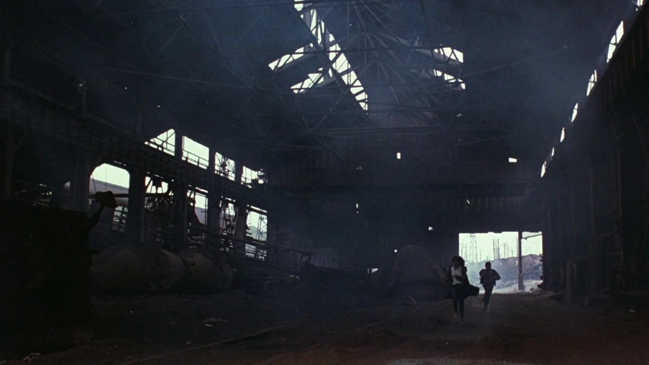 Flashdance (1983) movie still: wide — A vast, abandoned industrial steel mill with high ceilings and heavy structural metal beams. Two figures run…