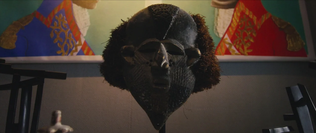 Da Sweet Blood of Jesus (2014) movie still: closeup — A dark, carved wooden mask with textured hair sits centered on a display stand. Two colorful…