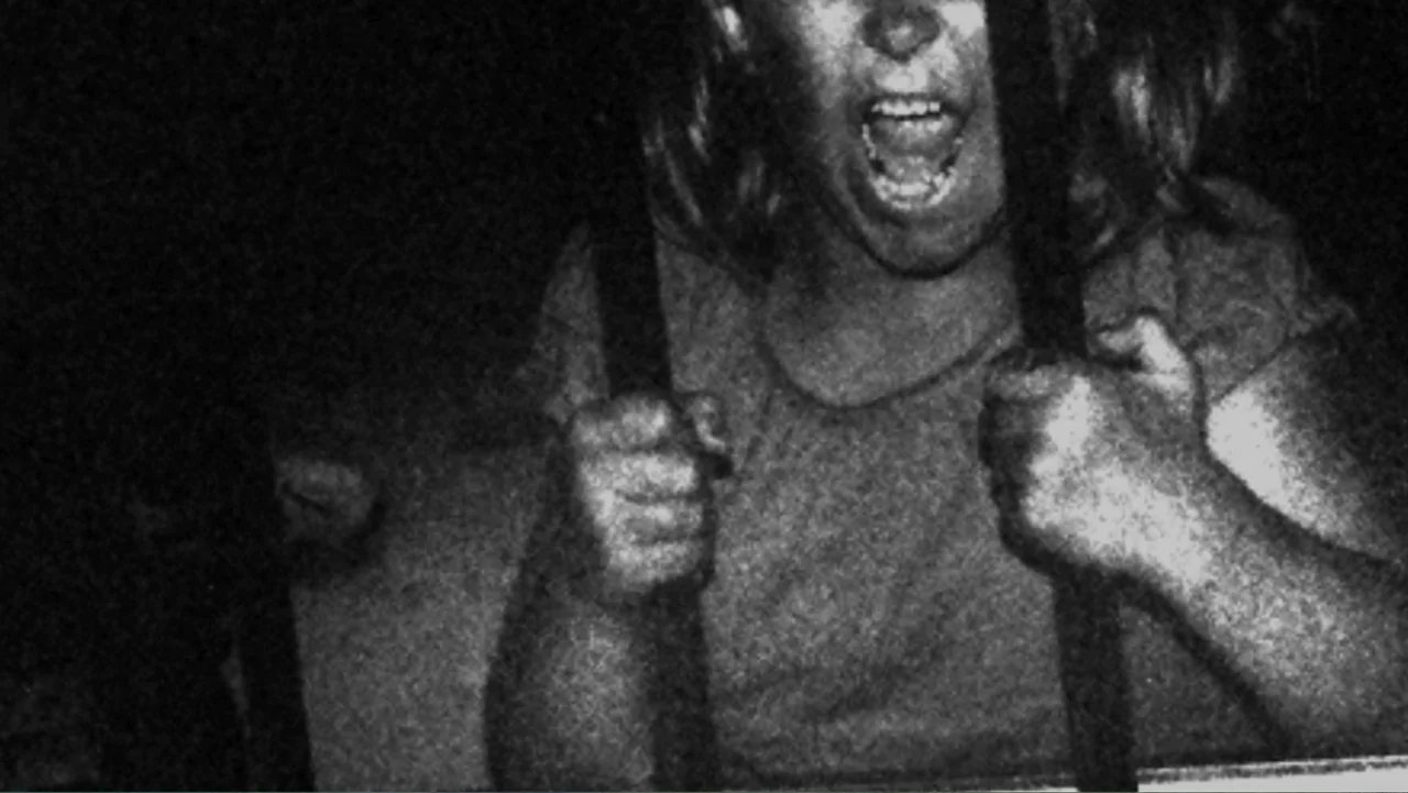 Savageland (2015) movie still: medium — A dark space defined by vertical metal bars in the immediate foreground. A young girl clutches the bars with…