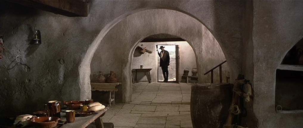 The Good, The Bad And The Ugly (1966) movie still: wide — A dimly lit room with thick, textured adobe walls and arched doorways. A man stands in the…