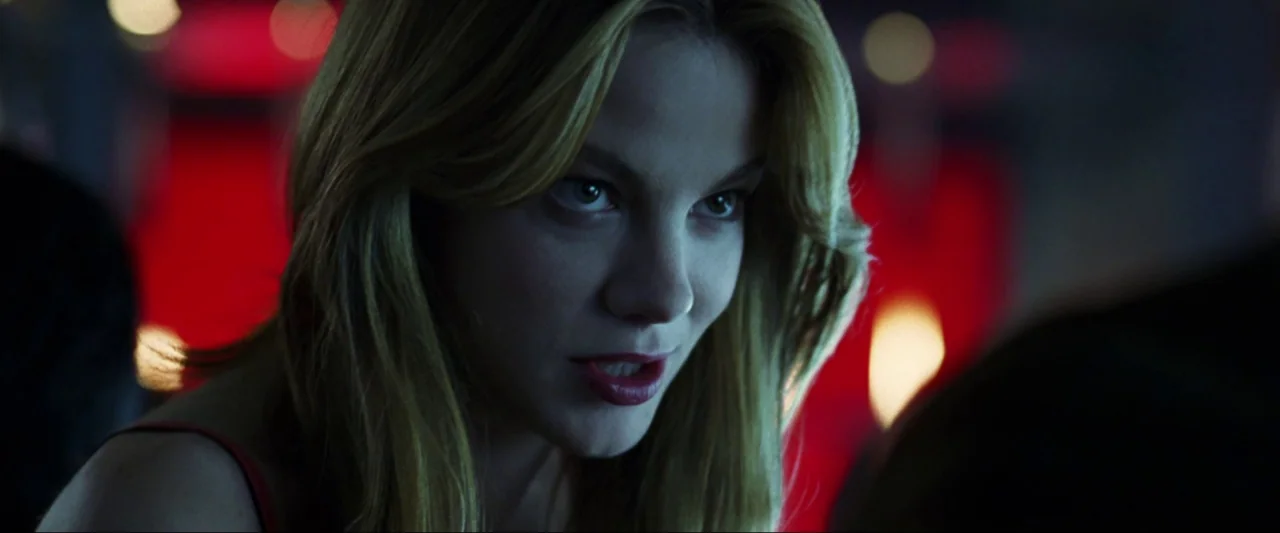 Kiss Kiss Bang Bang (2005) movie still: closeup — A dimly lit bar with red neon accents visible in the background bokeh. A woman with blonde hair…
