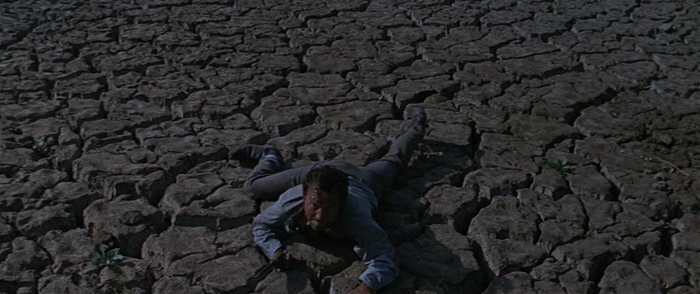 Flaming Star (1960) movie still: wide — An expanse of arid, deeply cracked mud stretches out under a clear, bright sky. A man lies prone on the…