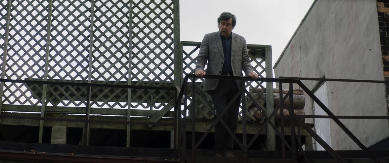 Greta movie still: fullbody — A man descends metal stairs outside a building featuring a wooden lattice screen wall. The sky is overcast and the…