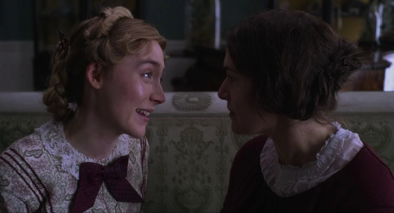Ammonite (2020) movie still: medium — Two women sit on a patterned fabric sofa inside a 19th-century parlor. They face one another while engaged in…
