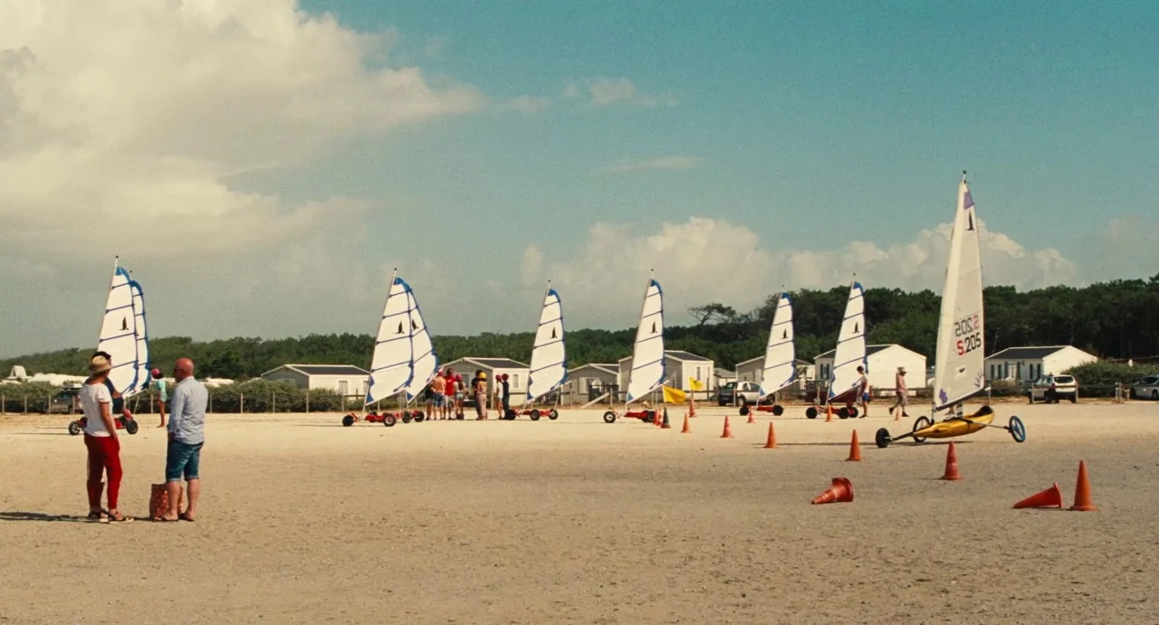 Ava (2017) movie still: wide — A flat sandy area features several land sailing carts with white sails positioned in rows. People gather near the…