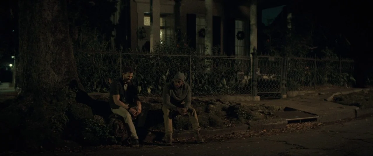 Synchronic (2019) movie still: wide — Two men sit on a dirt-covered curb beside a large tree at night. A wrought-iron fence separates them from a…