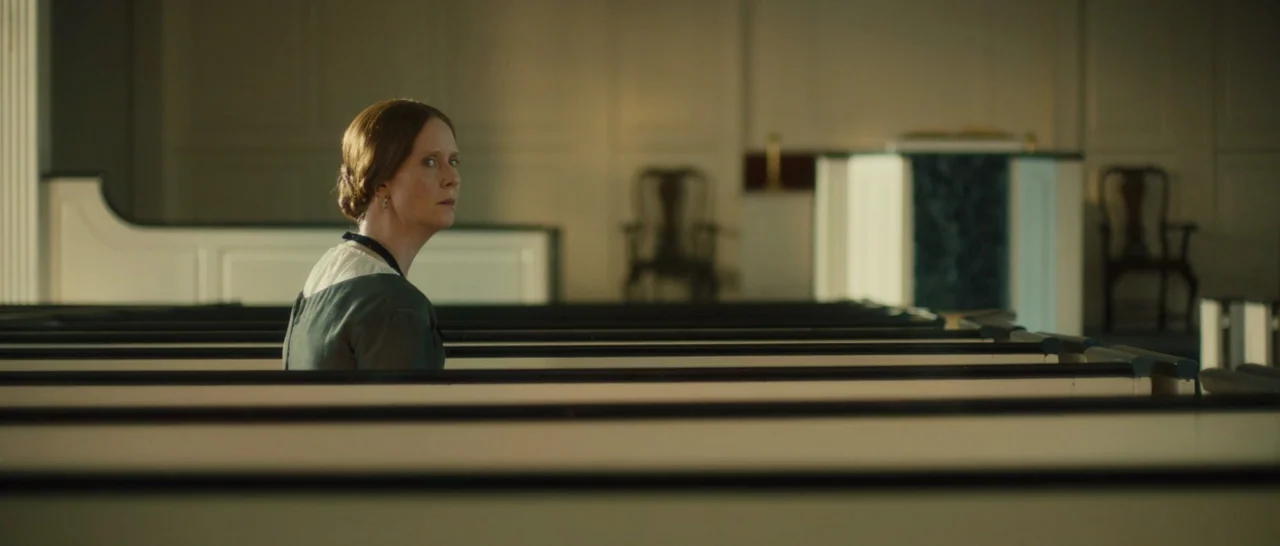 A Quiet Passion (2016) movie still: medium — An empty, minimalist church interior with rows of long, painted wooden pews. A woman sits alone in a…