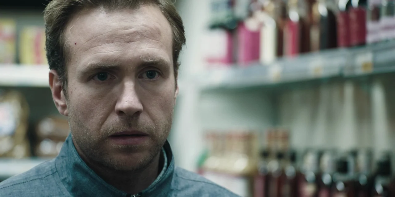 The Ritual (2017) movie still: closeup — A man stands in an aisle of a store with blurred shelves in the background. The man looks forward toward an…