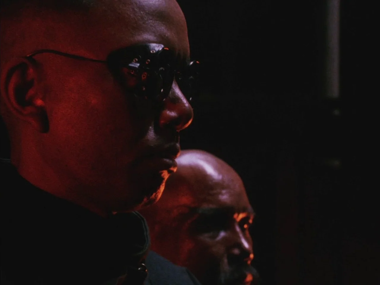 Tetsuo II: Body Hammer (1992) movie still: closeup — A dark, dimly lit environment where only the profiles of two men are visible. The two men sit…
