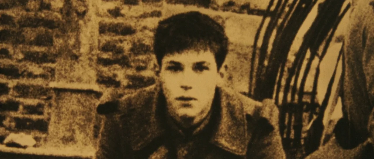 A Very Long Engagement (2004) movie still: medium — A teenage boy stands in front of a weathered brick wall. He stares forward while wearing a heavy…