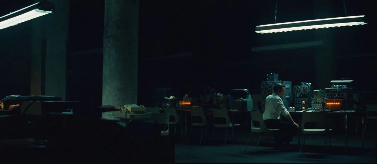 Seberg (2019) movie still: wide — A dark subterranean room contains rows of vintage audio monitoring equipment and concrete pillars. A man sits at a…