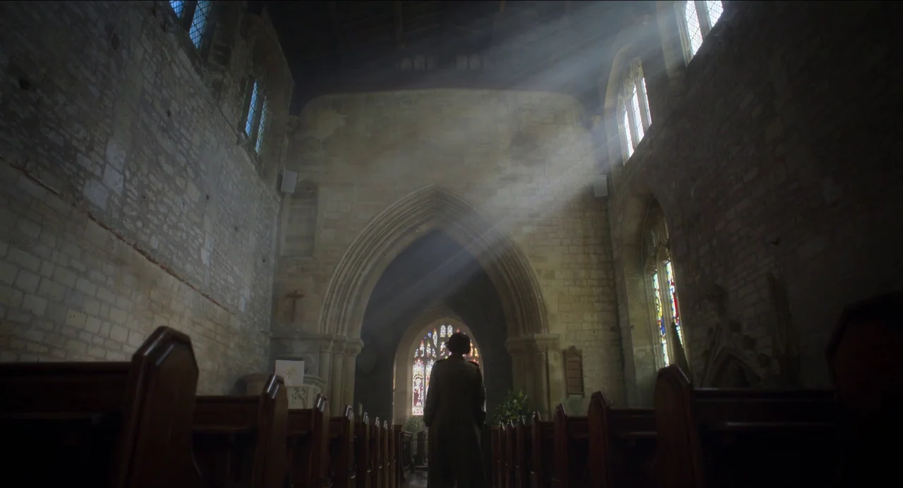 Men (2022) movie still: wide — An ancient stone church interior with high arched ceilings and rows of wooden pews. A single woman in a trench coat…