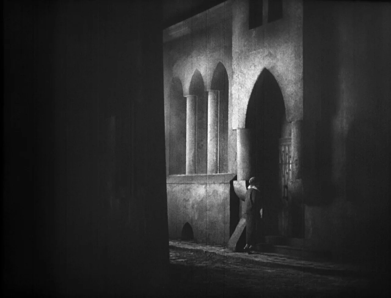 The Hands of Orlac (1924) movie still: wide — A narrow alleyway features stylized gothic stone architecture with arched windows and pillars. A…