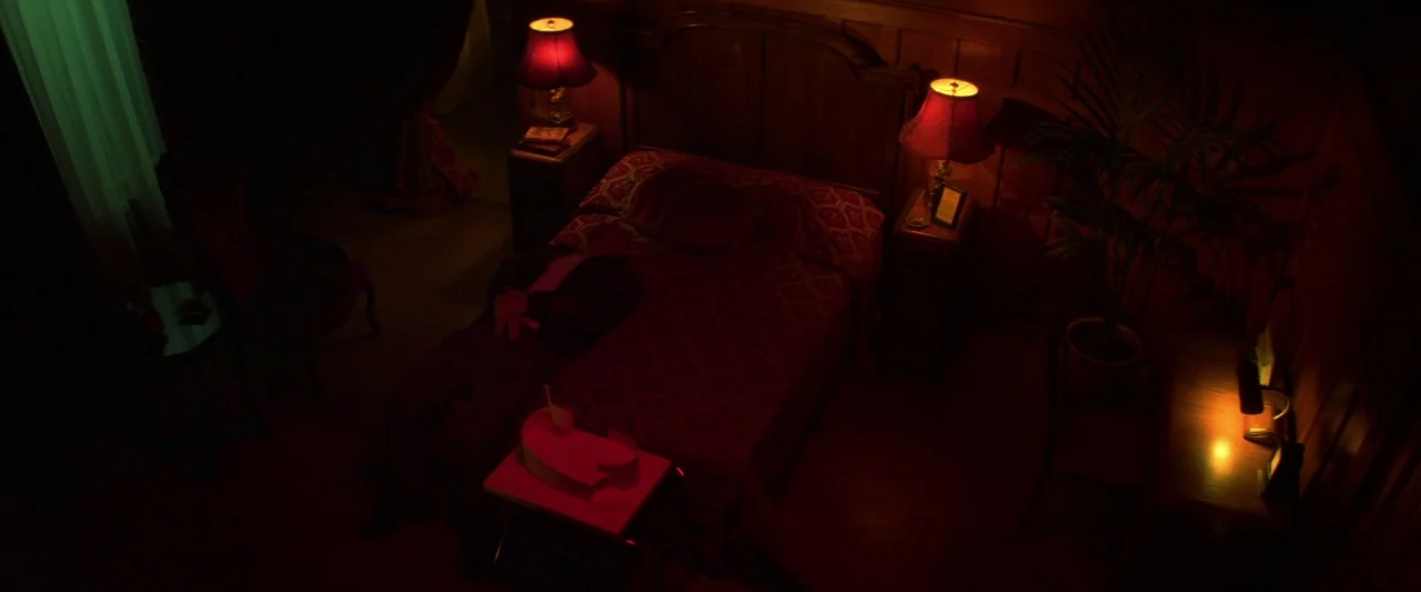 Zeroville (2019) movie still: wide — A dimly lit room with dark wood paneling and red decor elements. A person lies motionless on the bed under the…