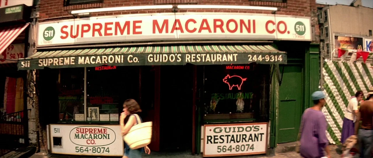 Léon: The Professional movie still: wide — A brick facade features a large sign for Supreme Macaroni Co. with green awnings. Passersby walk along…