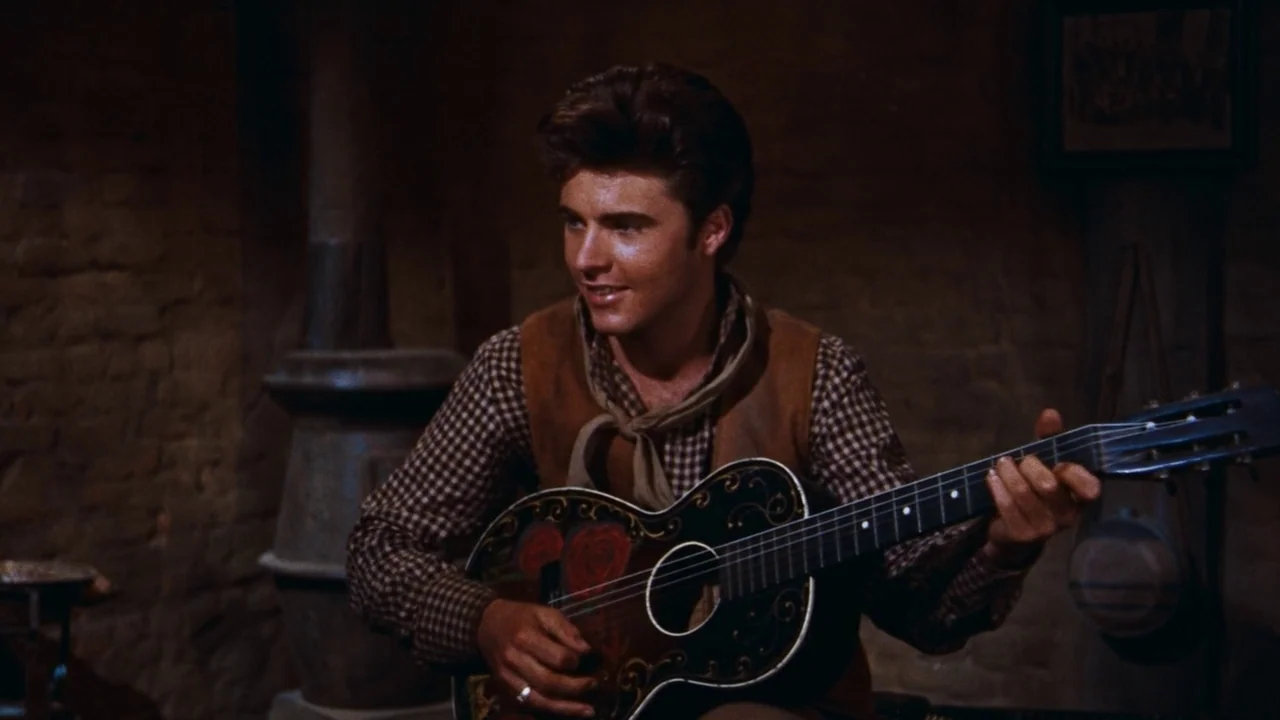 Rio Bravo (1959) movie still: medium — A dimly lit room with rough brick walls and an old cast-iron stove. A young man sits playing a…