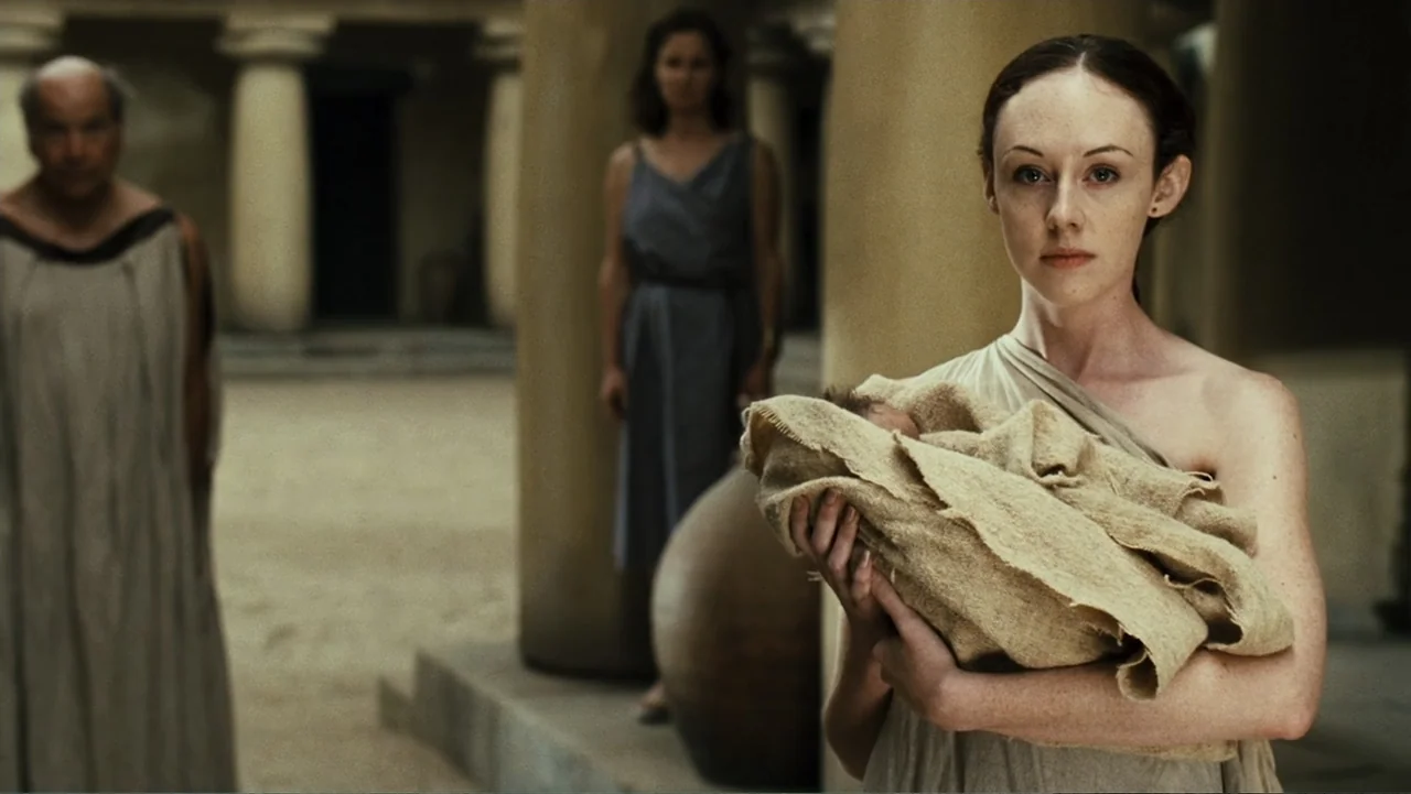 Meet the Spartans (2008) movie still: medium — An outdoor courtyard with stone columns and a sandy floor in an ancient setting. A young woman holds…