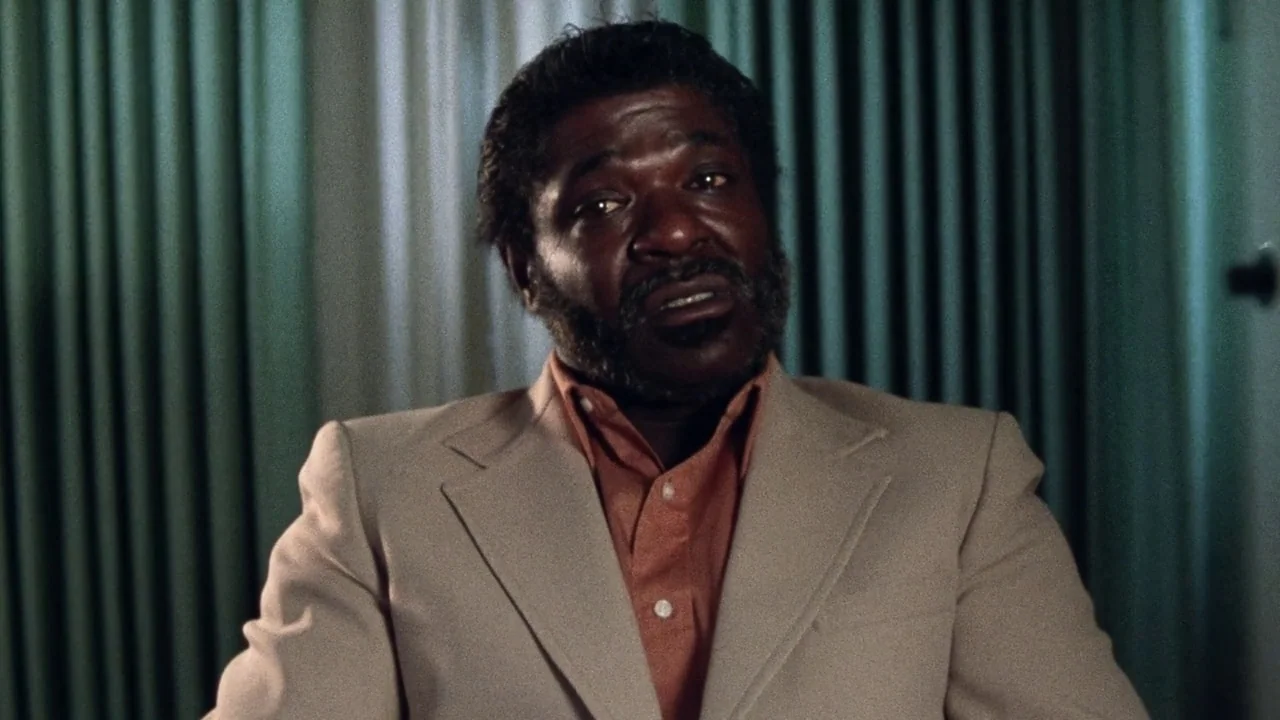 The Thin Blue Line (1988) movie still: medium — A man stands before pleated vertical blinds in a residential interior setting. He wears a beige…