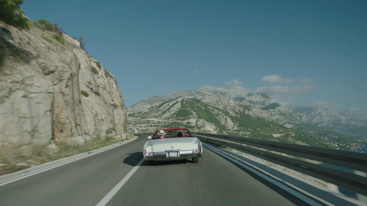 Infinity Pool (2023) movie still: wide — A white convertible travels along a paved coastal road carved into a steep mountainside. Rugged mountains…