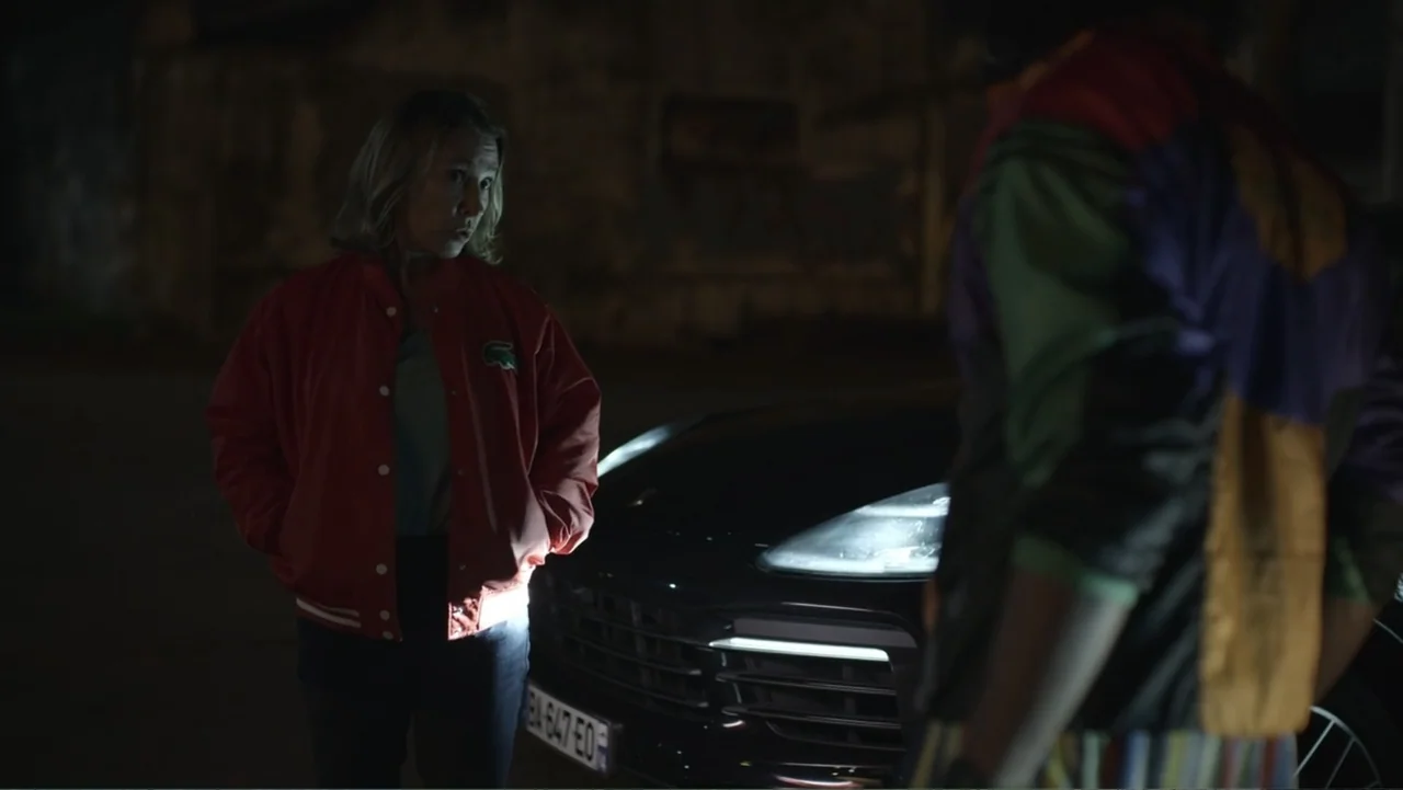 Unknown #1 movie still: medium — An exterior night setting features a black SUV parked on dark asphalt pavement. A woman in a red jacket stands…