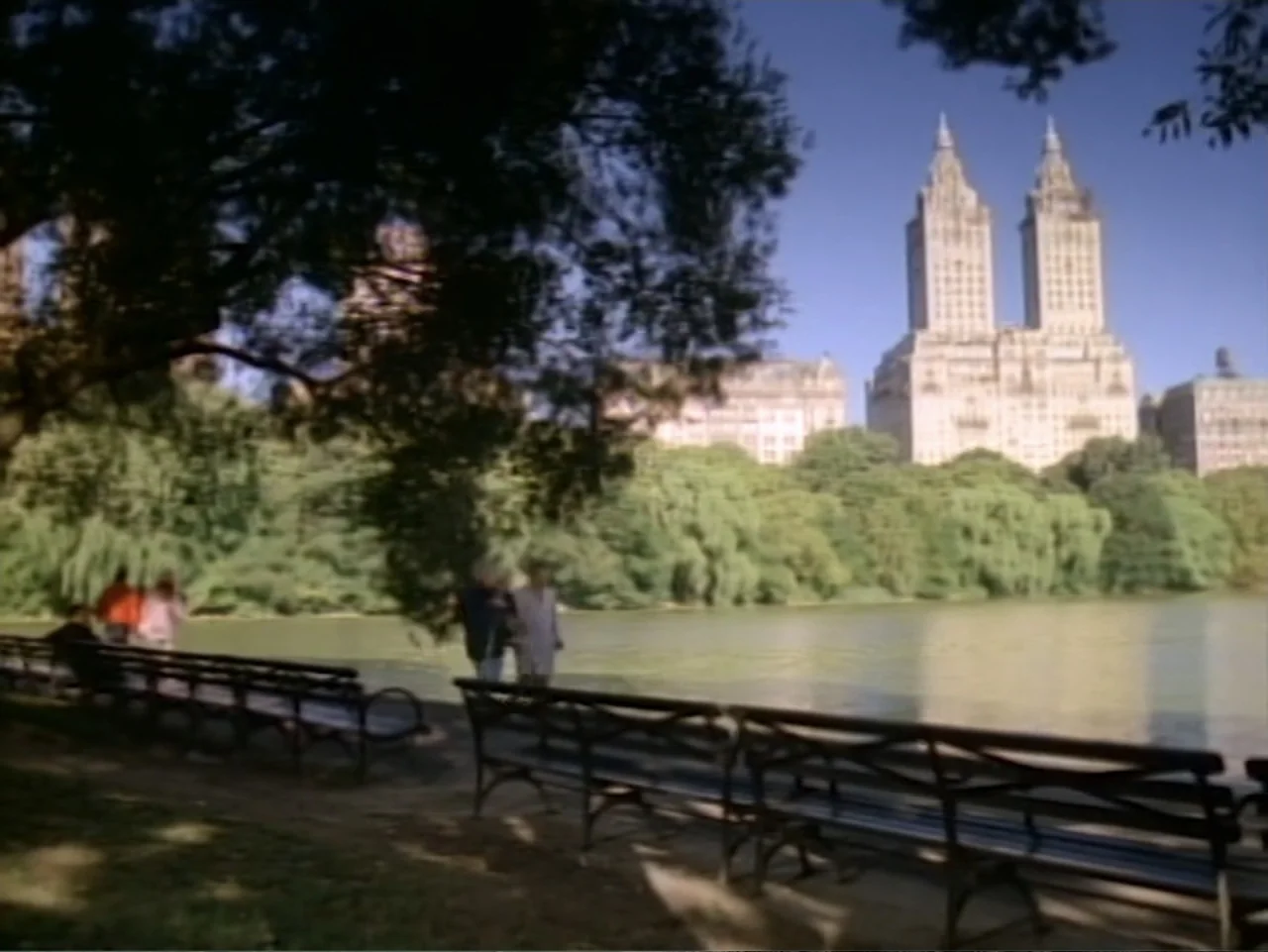My First Love (1988) movie still: wide — A public park features wooden benches lining a lake with twin towers rising behind trees. Distant figures…