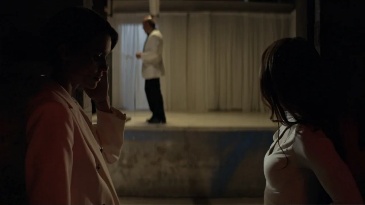 The Collective (2023) movie still: medium — A dark industrial space features an elevated stage with white curtains in the background. Two women…
