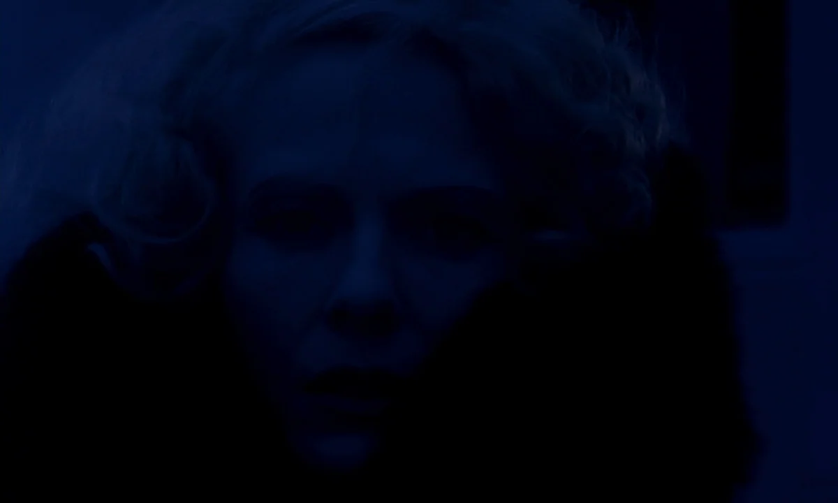 Daughters of Darkness (1971) movie still: closeup — A dark, dimly lit interior environment cast in heavy blue light. The subject looks directly…
