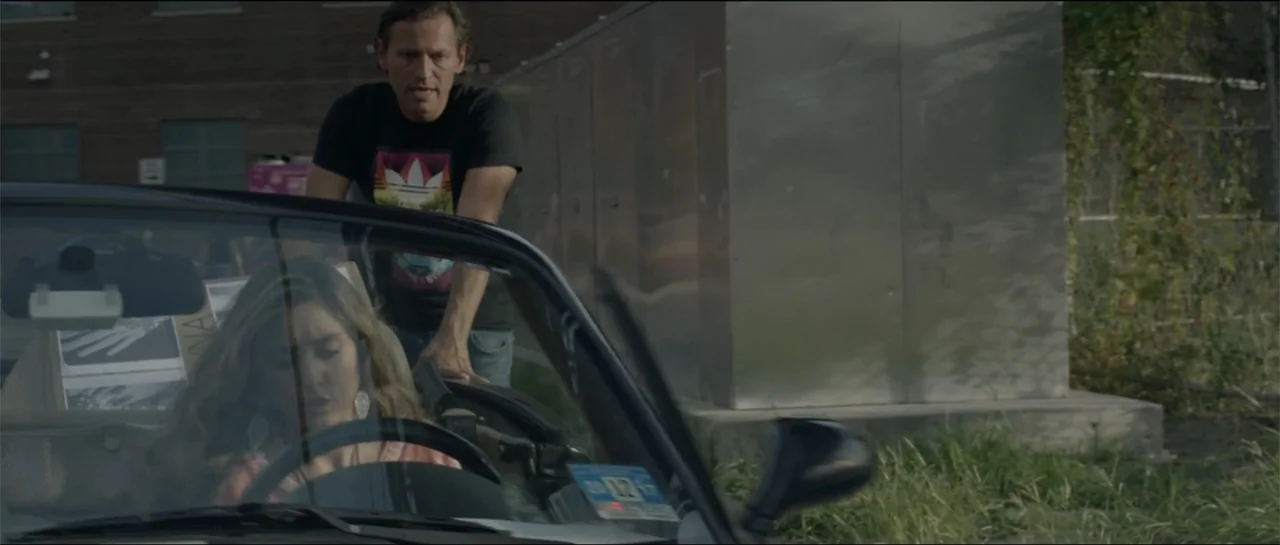 All She Wrote (2018) movie still: medium — A man stands beside a parked convertible car in a paved lot during daytime. A woman sits behind the…
