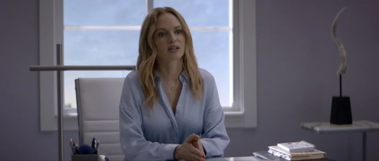 Suitable Flesh (2023) movie still: medium — A light-filled workspace with a white desk and a large window in the back. A blonde woman sits behind…