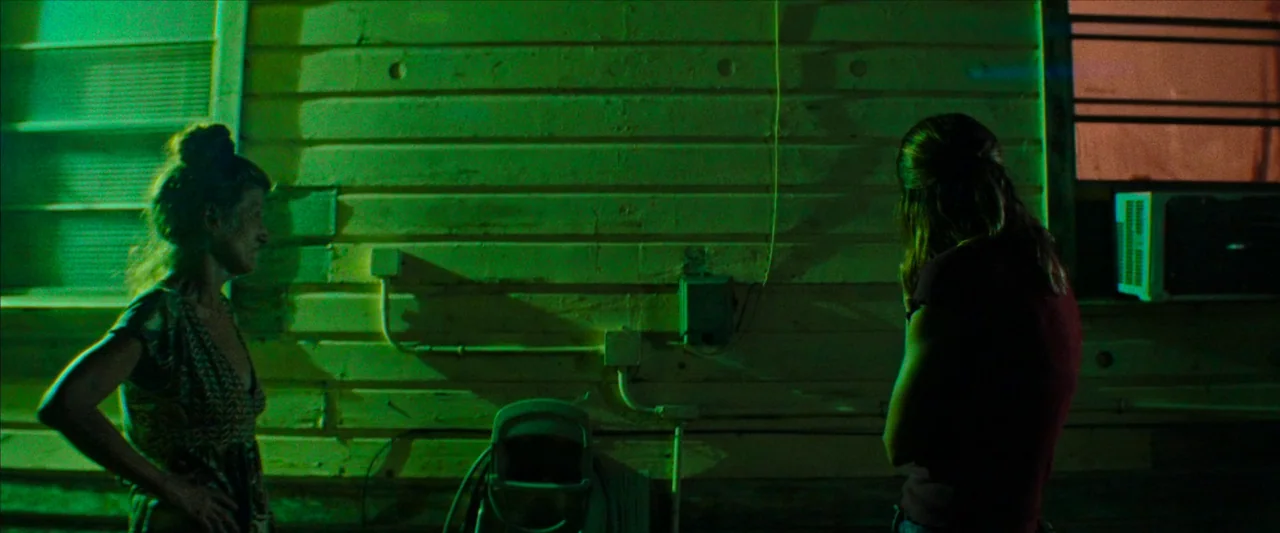 Red Rocket (2021) movie still: medium — The exterior of a house features horizontal wooden siding with a mounted utility box. Two women stand before…