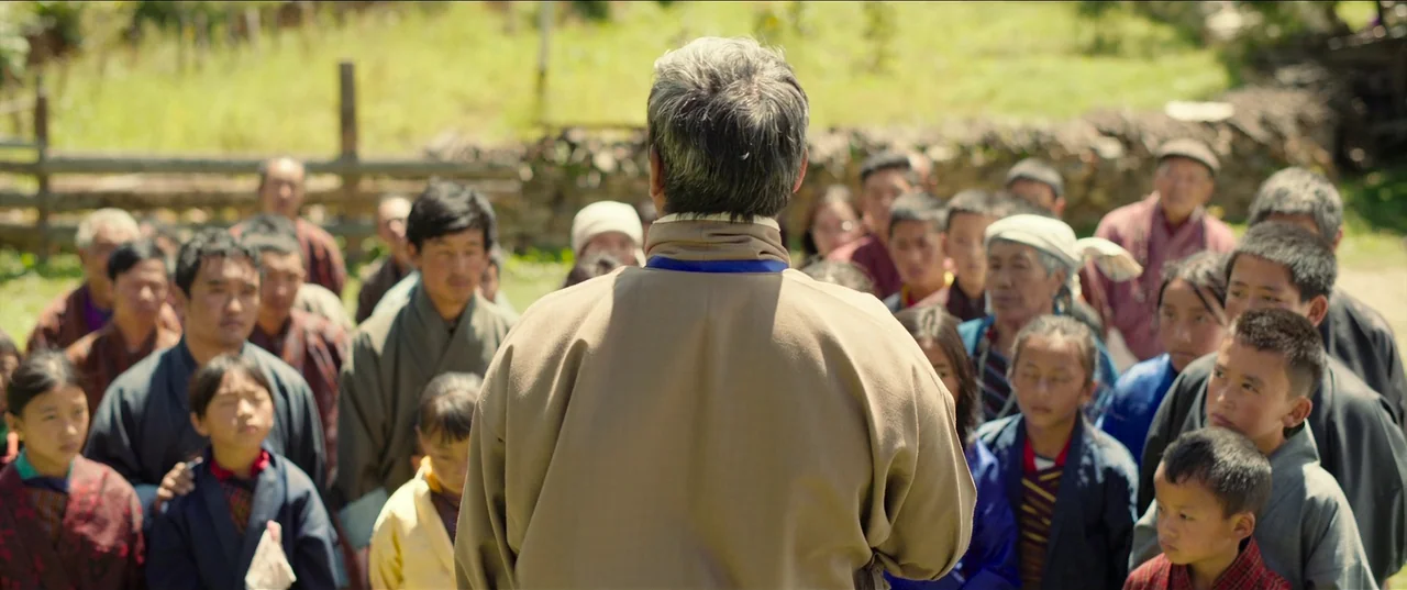 The Monk and The Gun (2023) movie still: medium — A crowd of people of various ages stands in a grassy outdoor location. The primary subject faces…