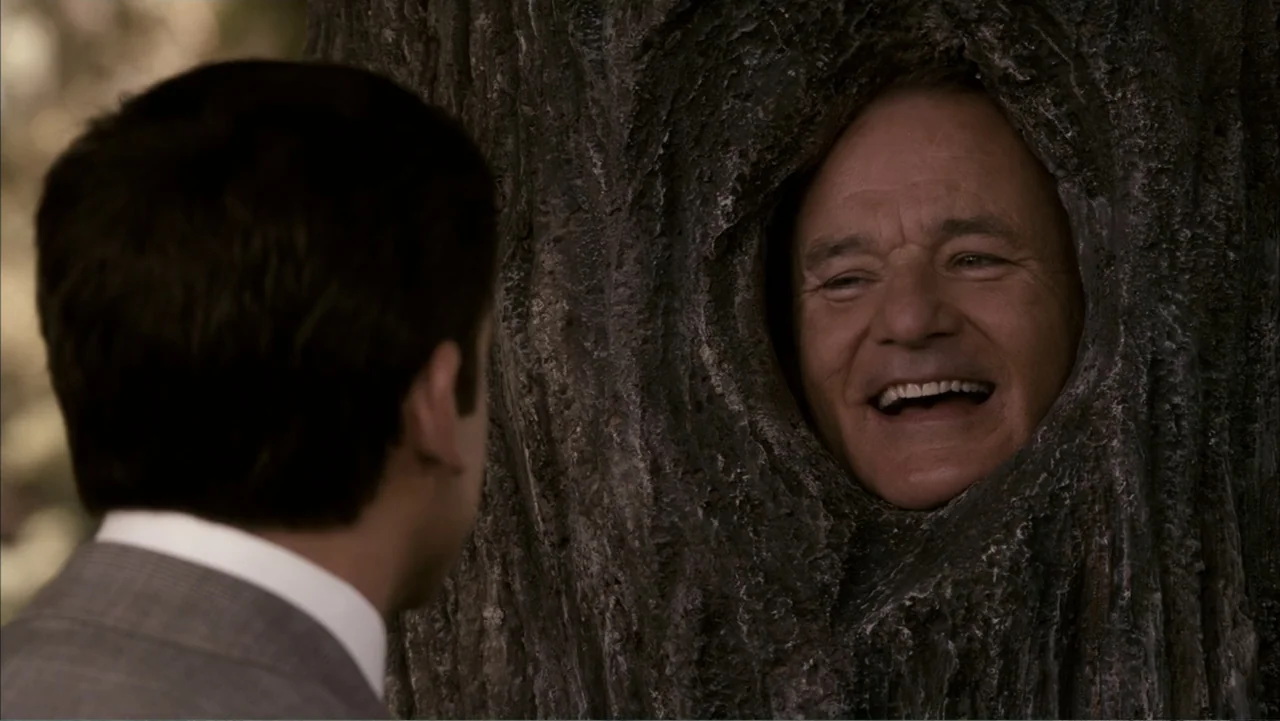 Get Smart (2008) movie still: medium — An outdoor park setting features a large textured tree with a hollow opening. A man appears to have his face…