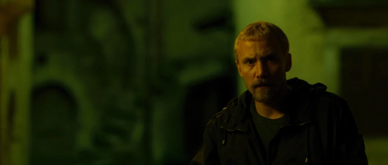 The American (2010) movie still: medium — A man stands in a dark, narrow stone alleyway at night. The man stares forward while illuminated by a…