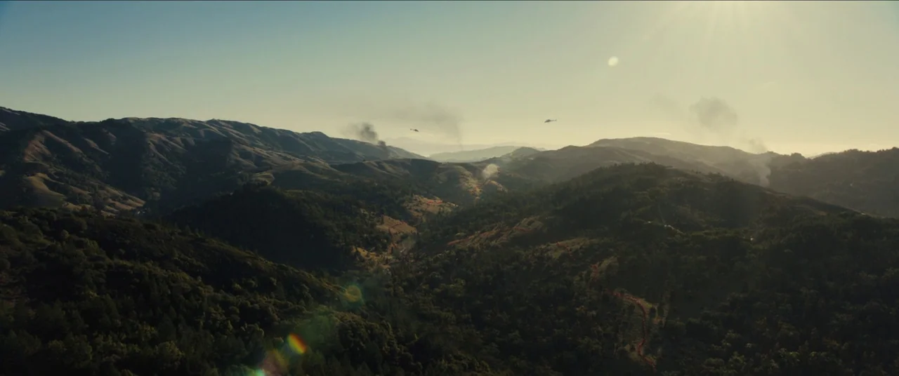 Us (2019) movie still: establishing — An aerial view of a sprawling mountain range covered in dense green forests. Several distant helicopters…