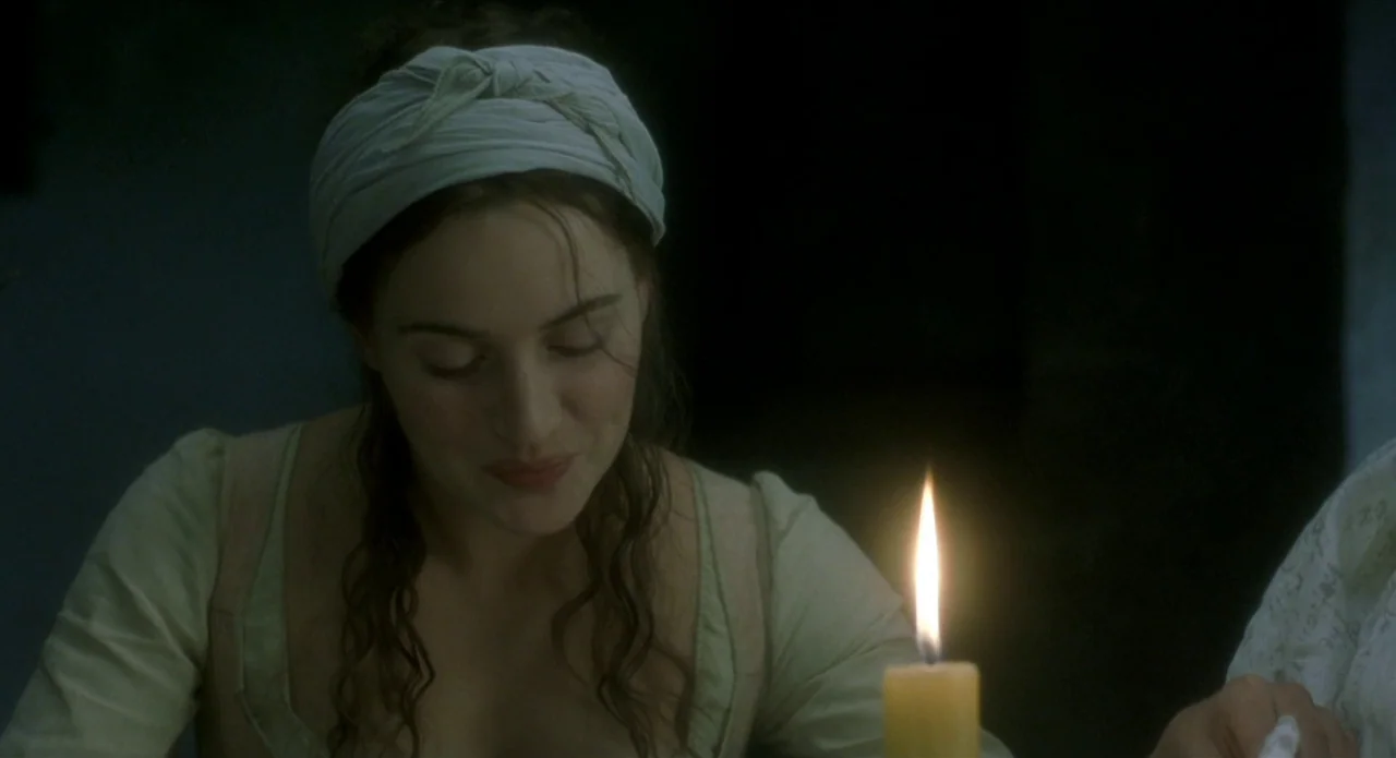 Quills (2000) movie still: closeup — A dim room illuminated primarily by the warm glow of a single candle. A woman with dark hair tied back sits at…
