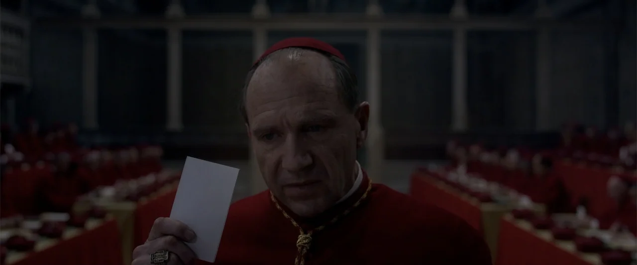 Conclave (2024) movie still: medium — A dimly lit hall features rows of seated figures in red robes. A man in clerical attire holds a white card…