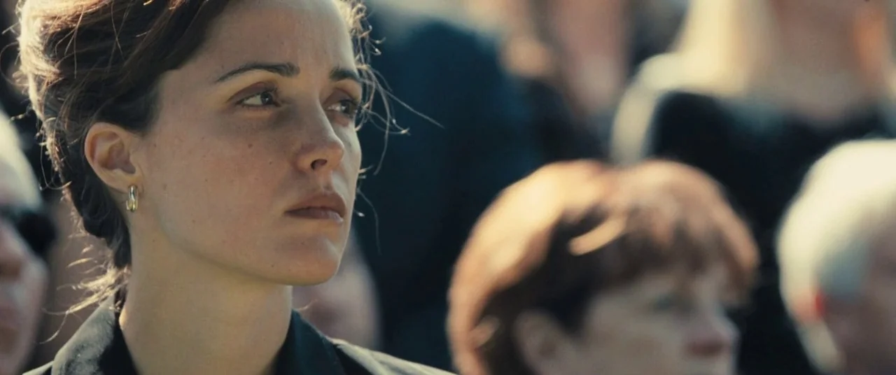 The Place Beyond The Pines (2012) movie still: closeup — A group of people stand outdoors in daylight, likely at a funeral or memorial. A woman in…